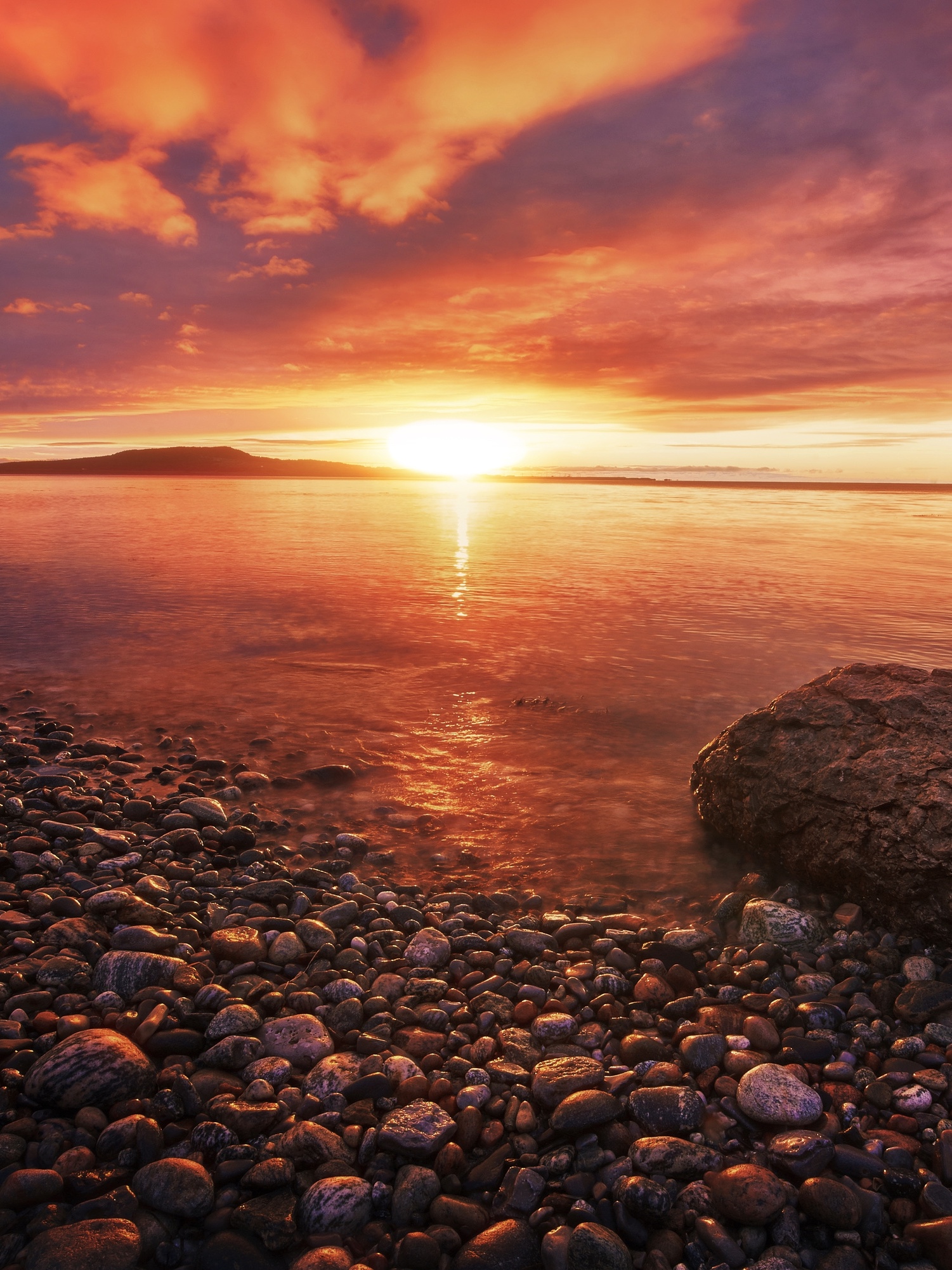 The sun setting on the shores of Shallow Bay in West Newfoundland. The shore is very rocky with large and small rocks. There is a large rock in the water on the shore. The sky is red, orange, and yellow. 