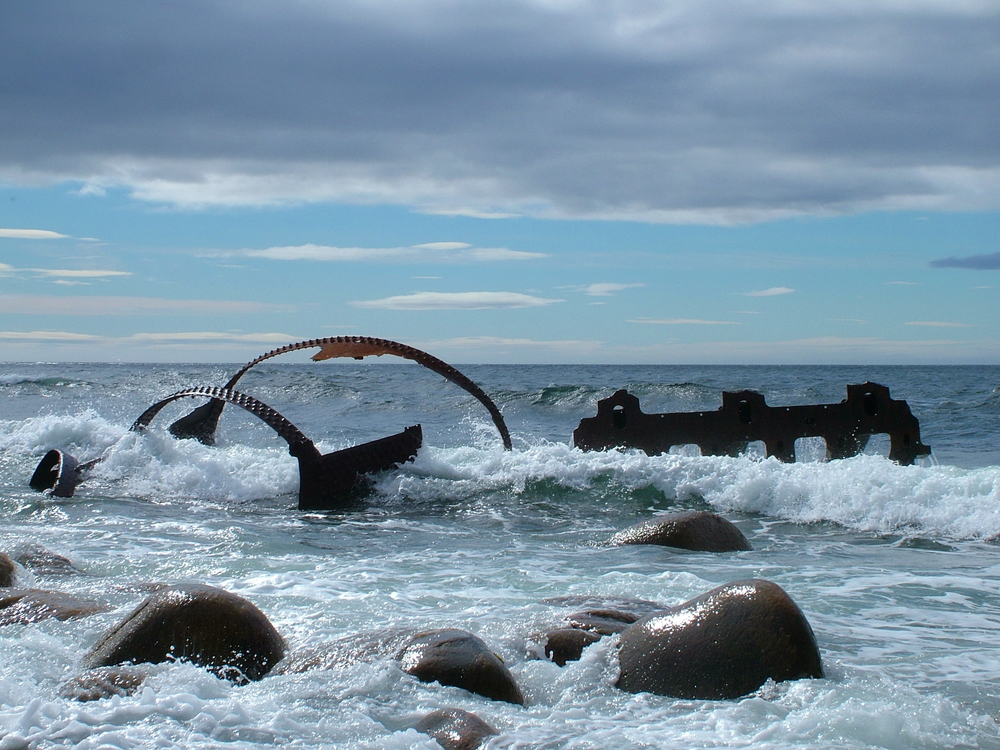 Remains of the SS Ethie shipwreck, one of the best stops on a West Newfoundland itinerary. You can see large rusted pieces of metal in the water with waves crashing against them. There are also large rocks in the water. 