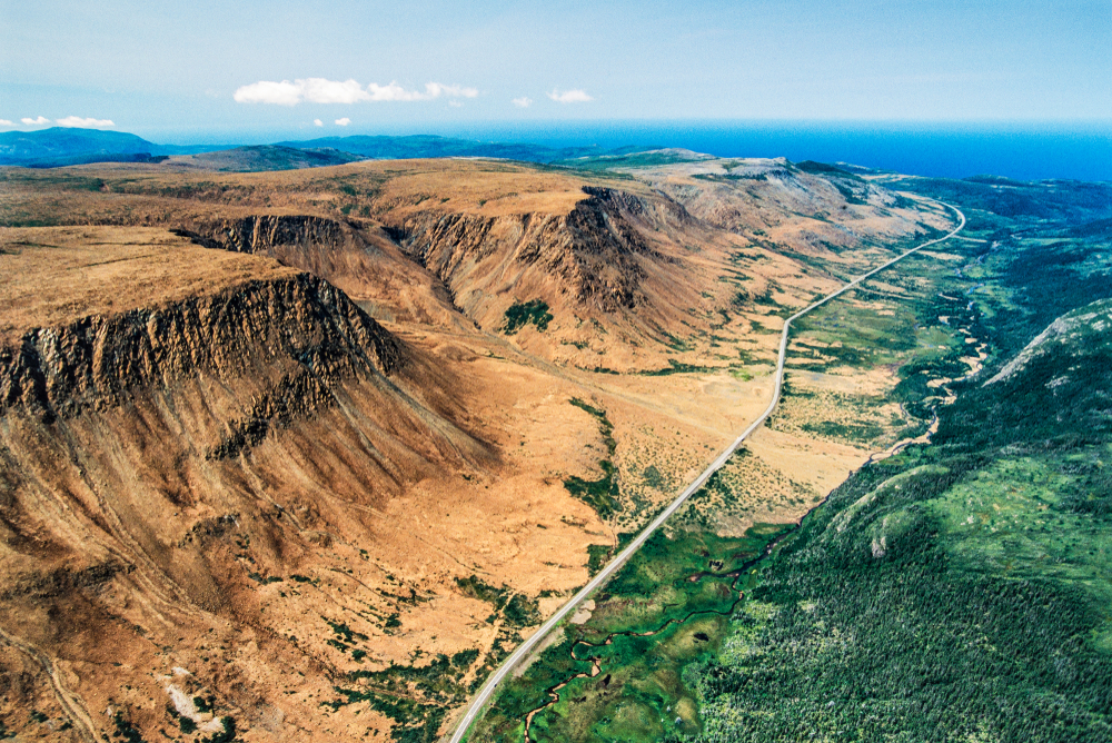 An aerial view of the Tablelands in Gros Morne National Park. It is a sandy looking landscape with large tall plateaus. In the valley there is a boardwalk going straight through it. On the other side of the boardwalk there is a lush green area. It is one of the best things to see on a West Newfoundland itinerary. 