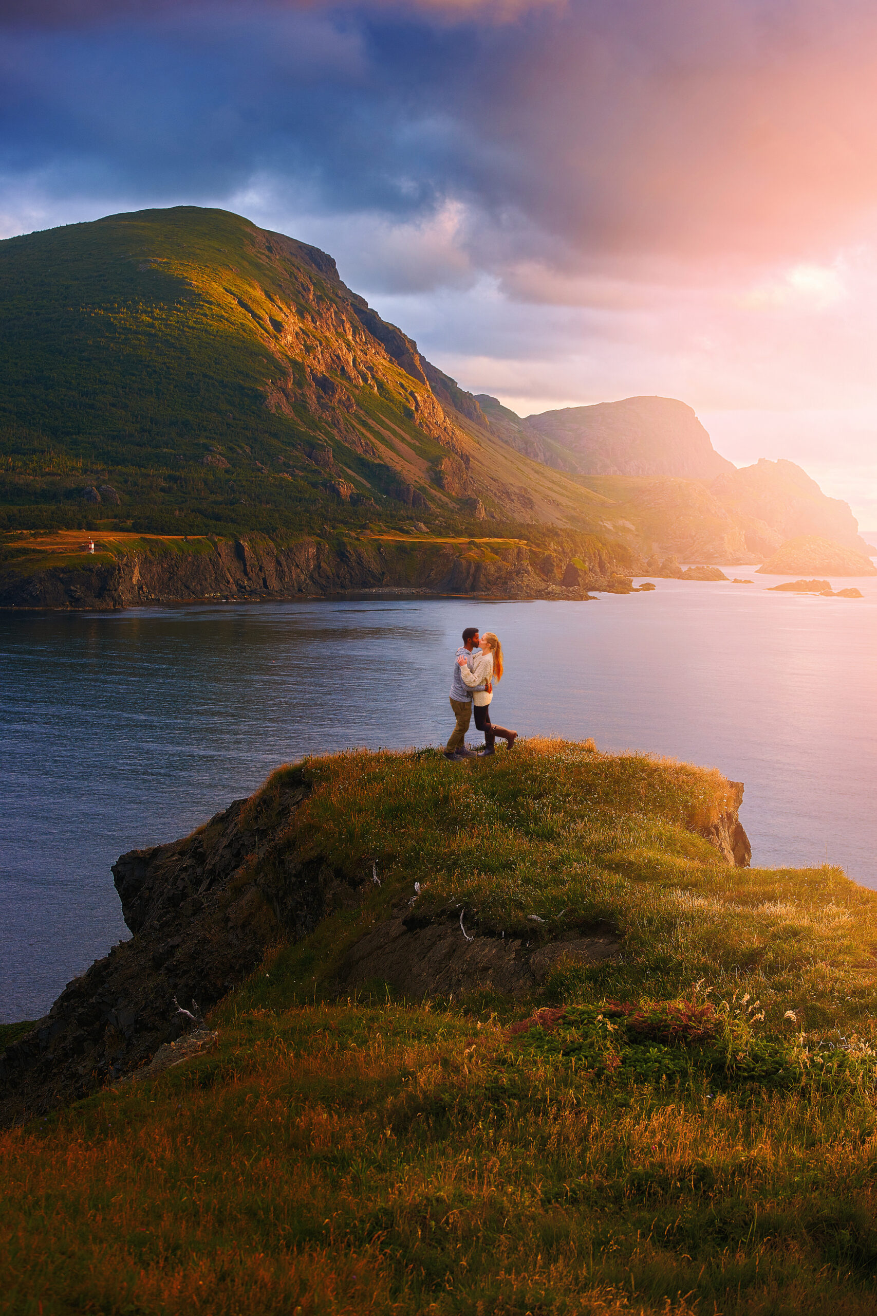 A couple kissing on the shore of the Trout River in Gros Morne National Park. There is a mountain range on the other side of the river. The sun is starting to set. It is one of the best stops on a West Newfoundland itinerary. 