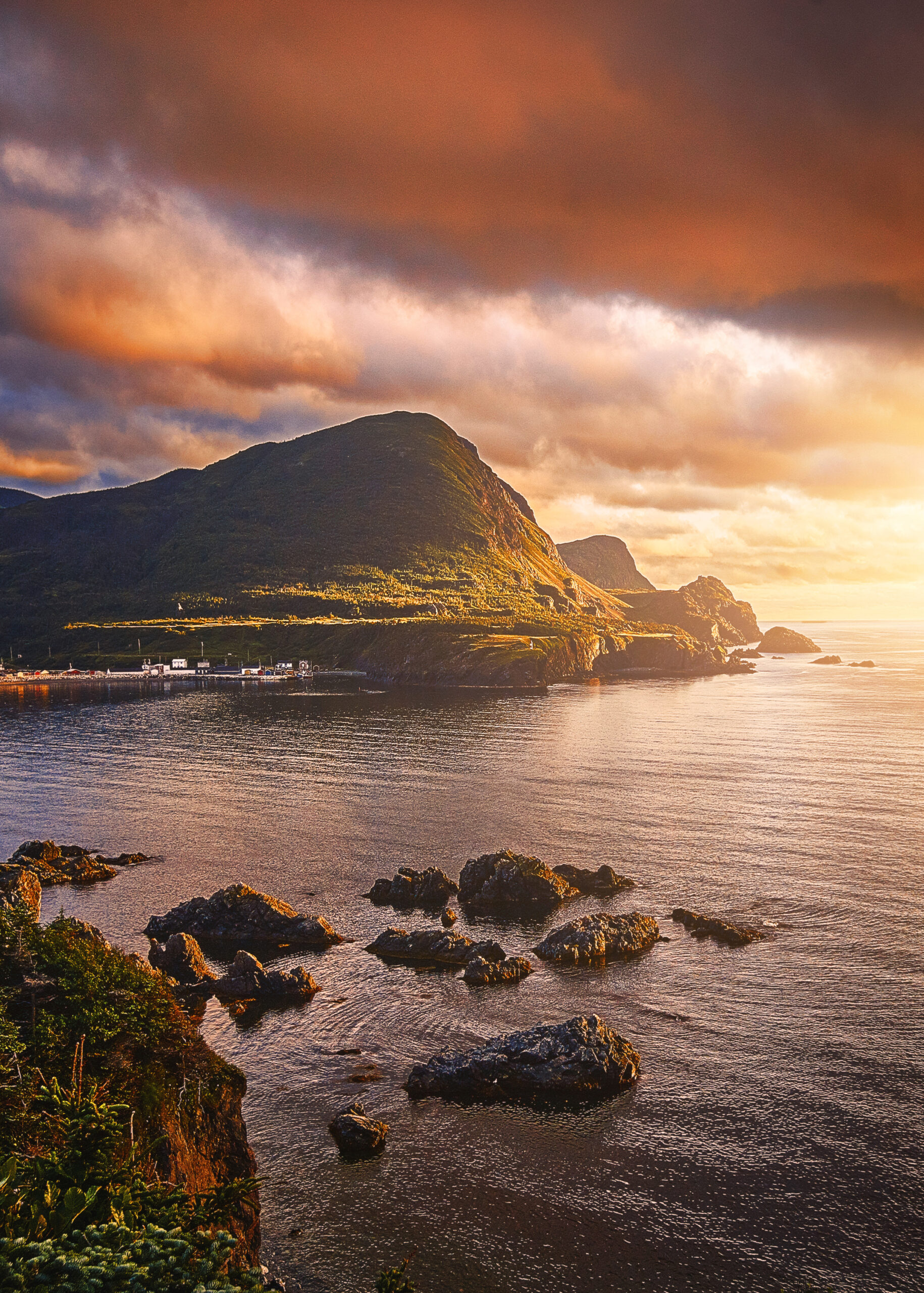 The view of a bay in West Newfoundland at sunset. There is a large hilly area, rocks in the water, and across the water is a dock. 