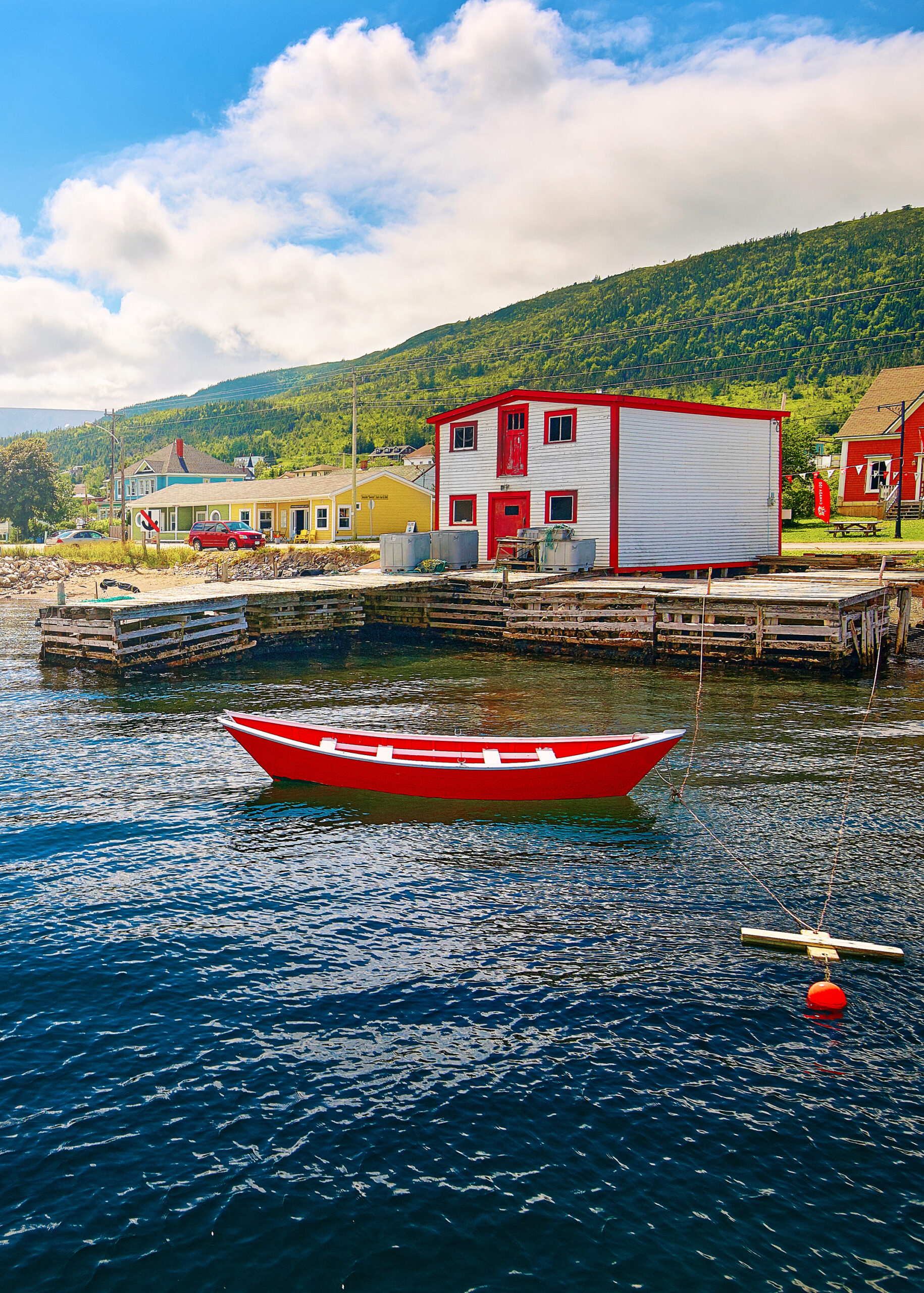 A white and red building on the shore of a bay in Woody Point. There is a red row boat with white trim floating in the water. You can see other colorful buildings that are yellow, blue, and red, in the back ground. There is a hill covered in trees. It is one of the best stops on a West Newfoundland itinerary. 