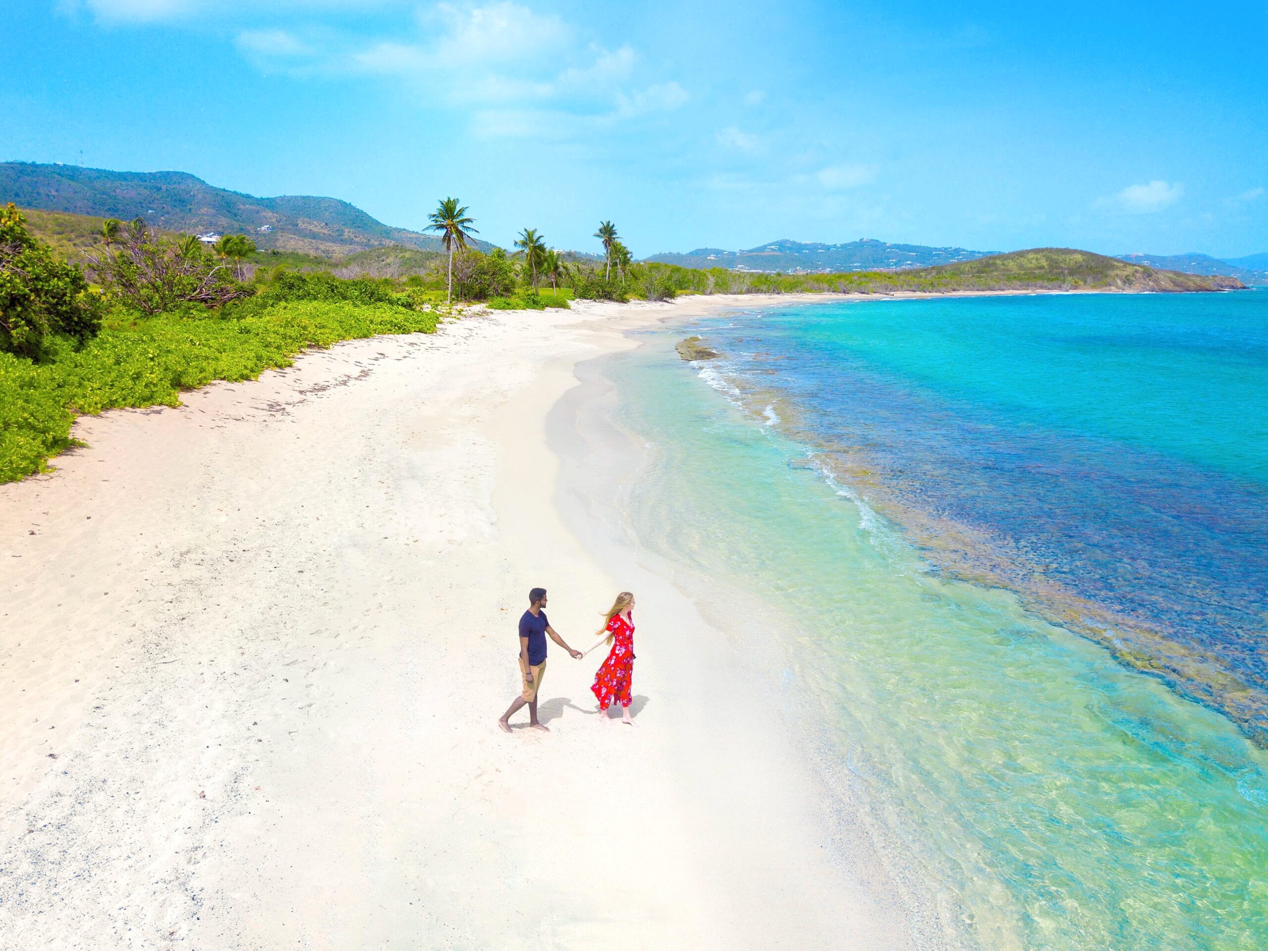 A man and a woman standing on a white sand beach looking towards the ocean. It is an aerial view and you can see a lush green island in the distance. One of the best islands in the USA.