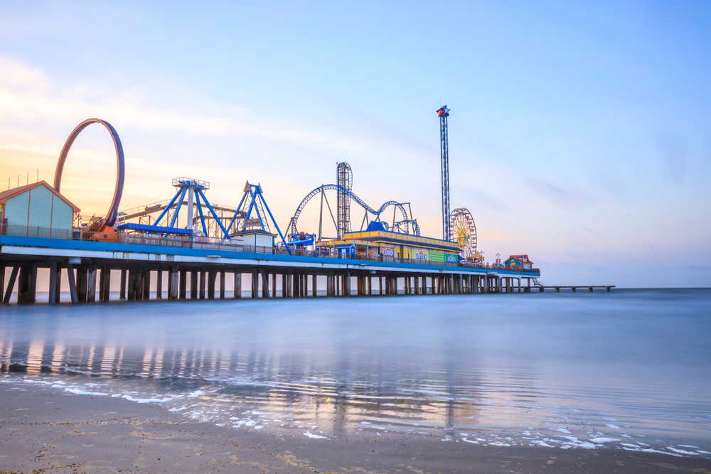 The Galveston Pier with amusement park rides on it as the sun is setting. The water is calm and a pale blue. 