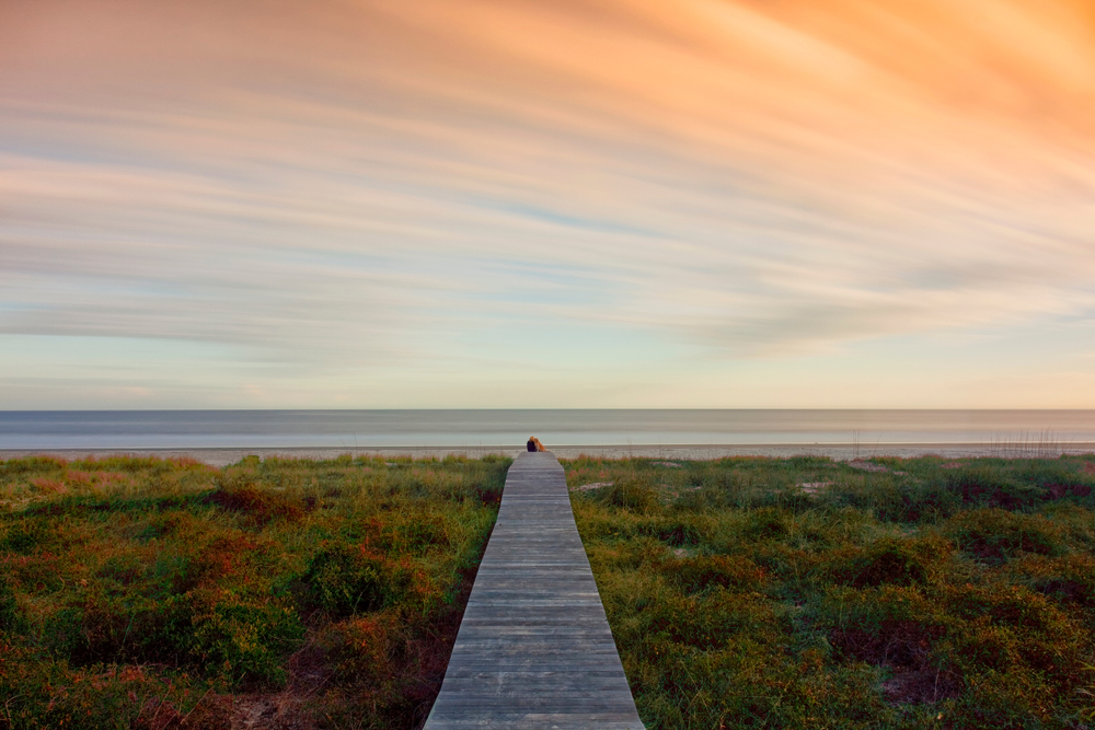 Looking down a long wooden pier that leads to the shore of the beach at Hilton Head Island South Carolina. The pier is surrounded by marsh and the sun is setting. 