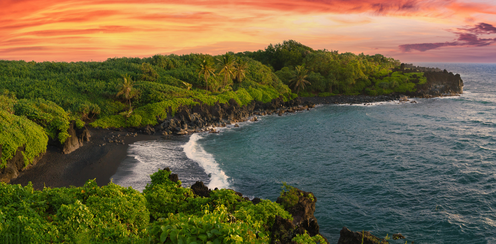 A view of a black sand beach in Maui. You can see a lush rainforest, the beach, and the bright blue water. The sun is setting. 