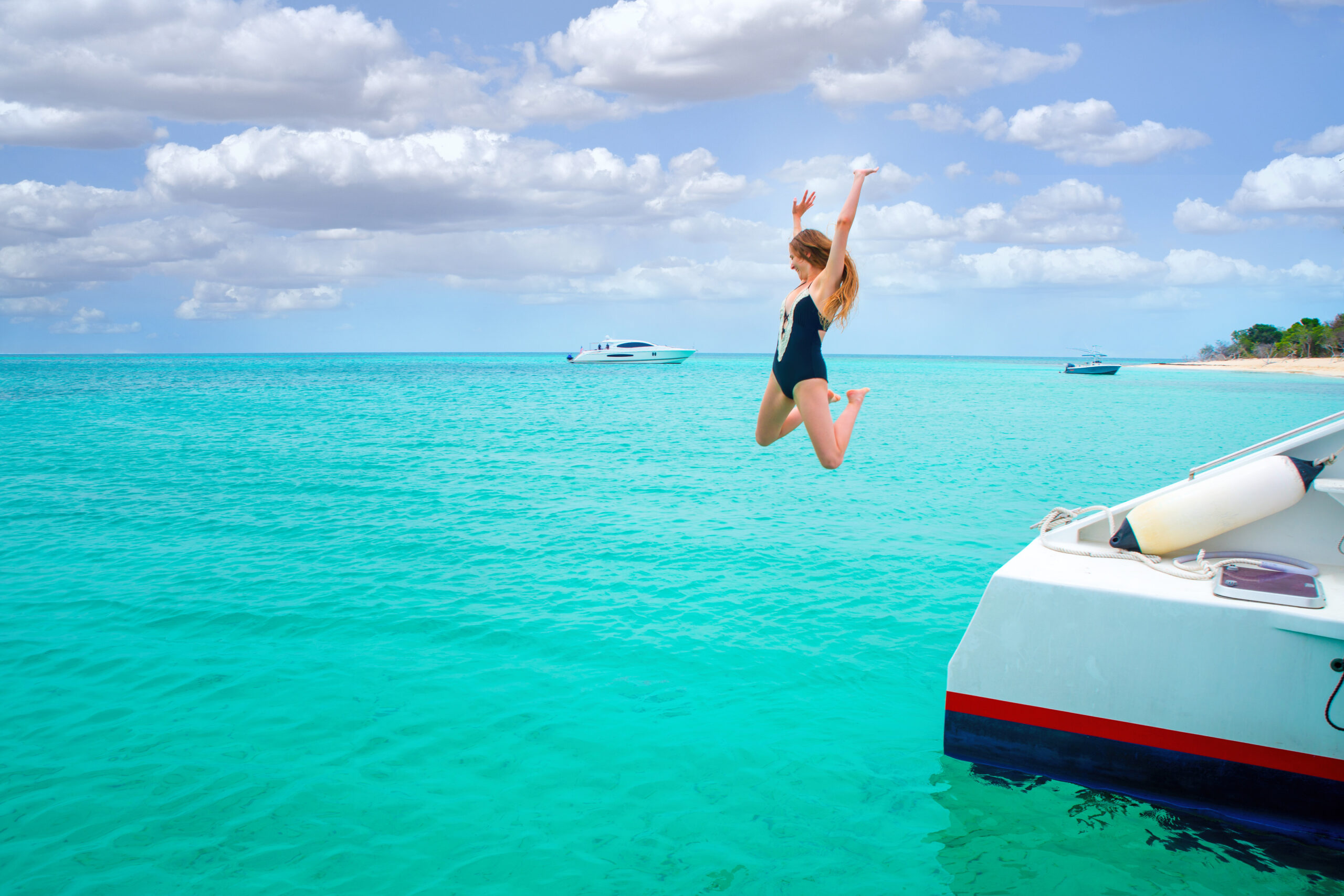 A woman in a black bathing suit jumping into crystal blue water at one of the islands in the USA. In the background you can see boats and a bit of sandy beach. 