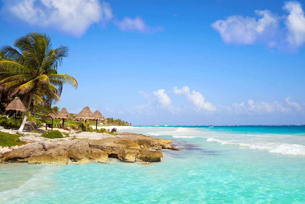 A beach in Mexico with crystal blue water, palm trees, white sand, and little huts on the shore. 