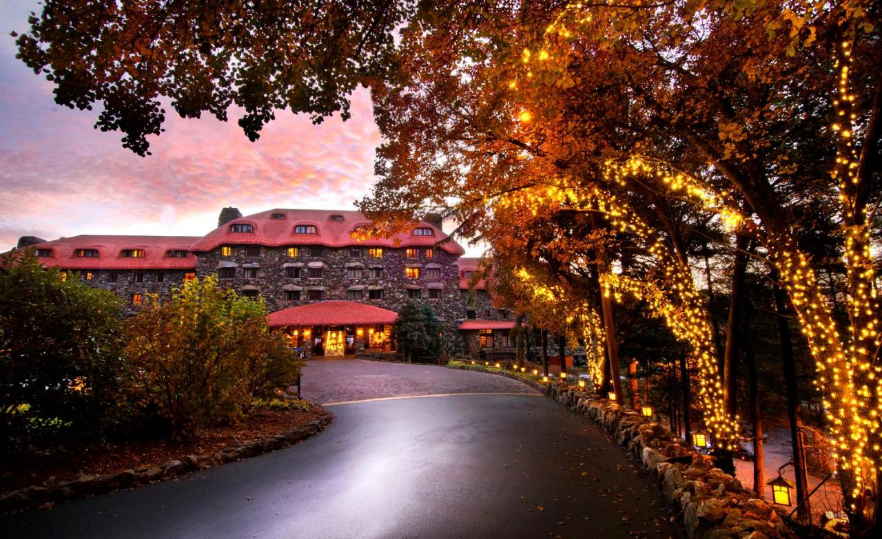 The front of a large stone luxury hotel as the sun is setting. There is a wide driveway with trees that have twinkle lights on them leading up to the hotel. 