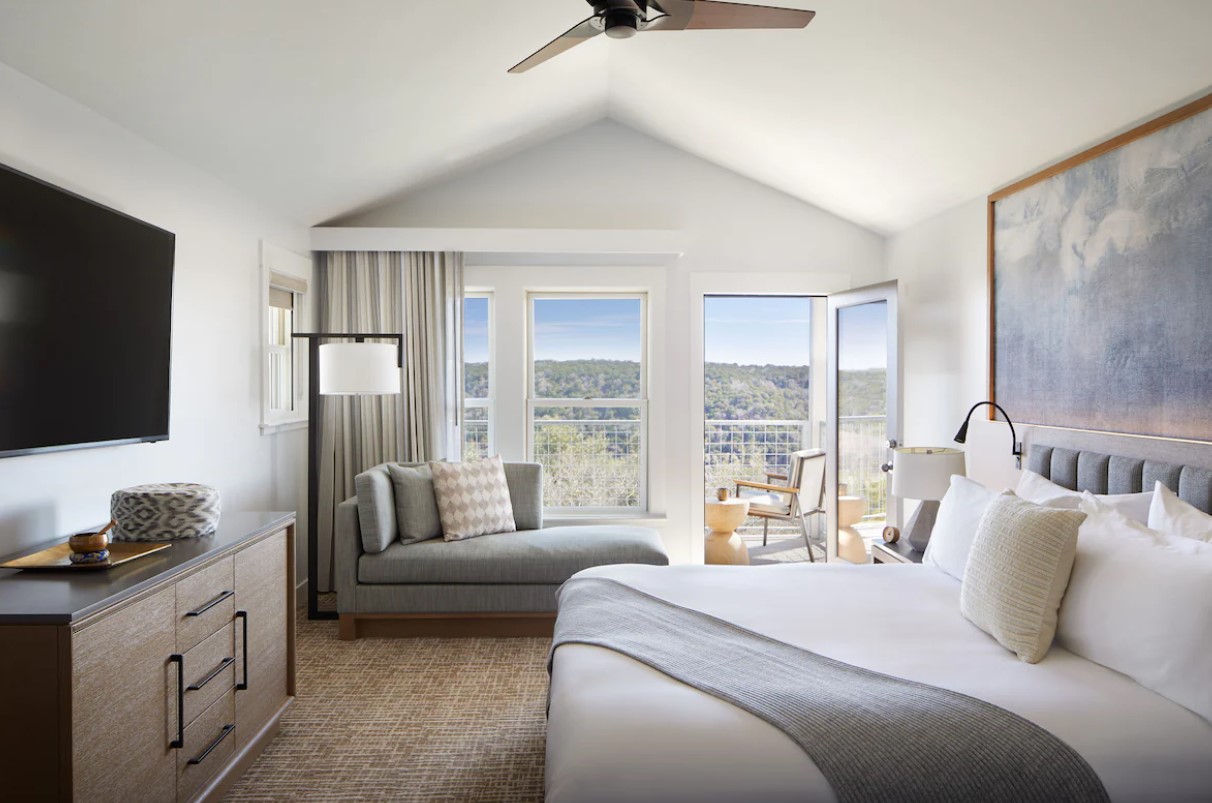 The view of a hotel room that has a king-sized bed, a small settee, and a balcony that looks out onto hills. 