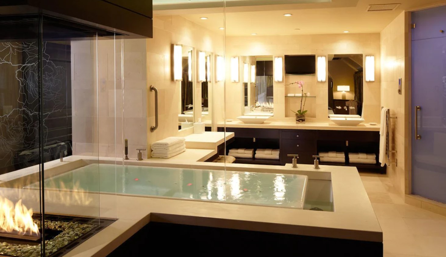 A large luxurious bathroom with a glass walled fireplace, a large soaking tub, a standing shower, and a double vanity. 