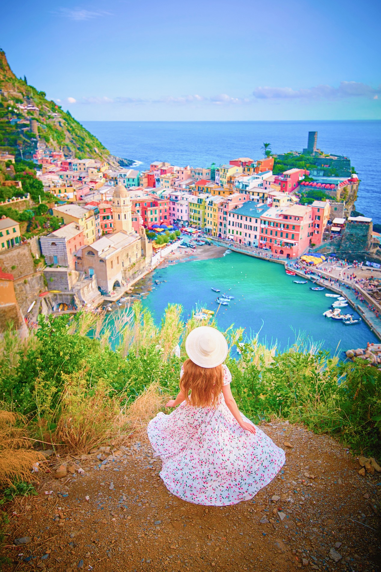 Woman in a floral dress looks down at one of the Cinque Terre villages and the harbor.