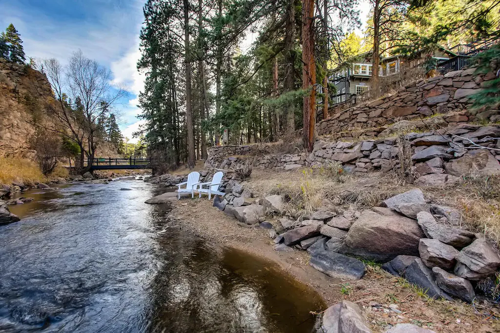 A cabin sits on rocky outcrop above a creek. Two chairs are by the creek. 