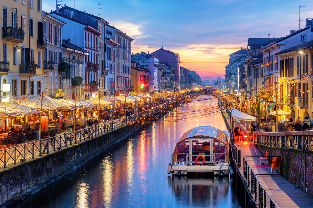 Sunset over the Navigli Grand Canal with a boat in the water, string lights, and busy restaurants.