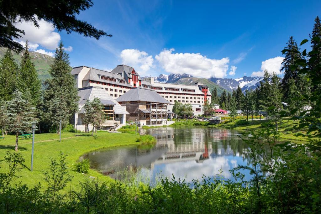 A large white lodge with a lake in front of it and large mountains behind it on a sunny day