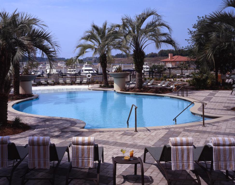 Photo showing a pool surrounded by sunbeds and palm tress with a harbor in the background. 