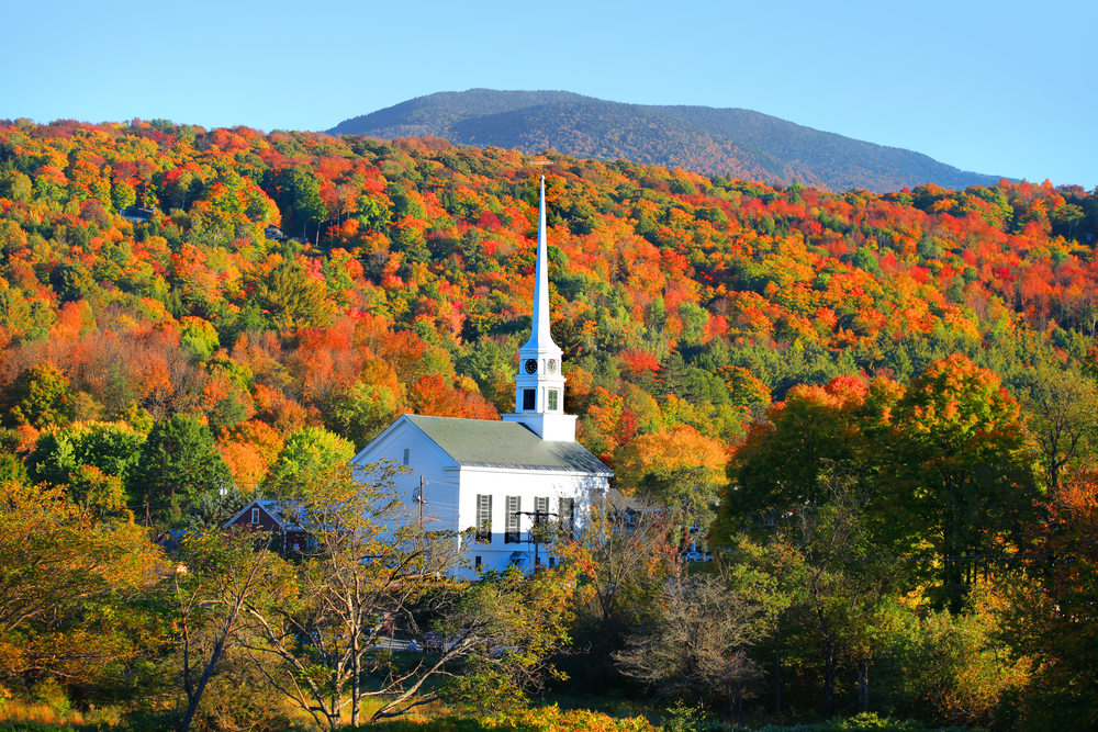 White church with tall steeple stands among rolling hills of fall foliage.