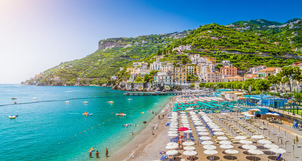 Dozens of beach umbrellas are set up in rows on a sunny beach with green hills in the background on the Amalfi Coast which has some of the best beaches in Italy.