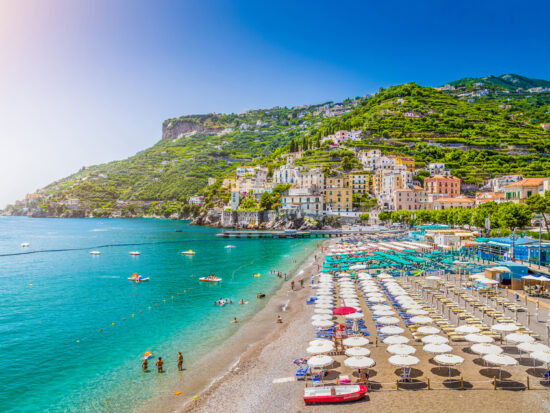 Dozens of beach umbrellas are set up in rows on a sunny beach with green hills in the background on the Amalfi Coast which has some of the best beaches in Italy.
