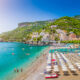 Dozens of beach umbrellas are set up in rows on a sunny beach with green hills in the background on the Amalfi Coast which has some of the best beaches in Italy.