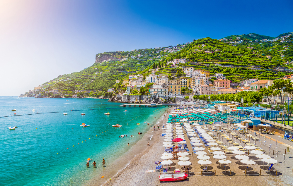 Dozens of beach umbrellas are set up in rows on a sunny beach with green hills in the background on the Amalfi Coast which has some of the best beaches in Italy.