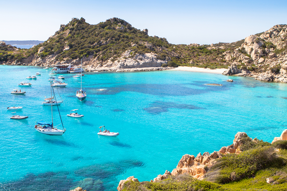 Small boats dot a turquoise cove with a small sandy beach on an uninhabited island that has the best beaches in Italy.