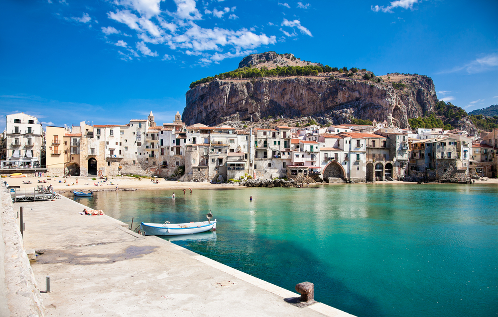 Light-colored buildings surround the quiet blue bay of Cefalu, one of the best beaches in Italy.