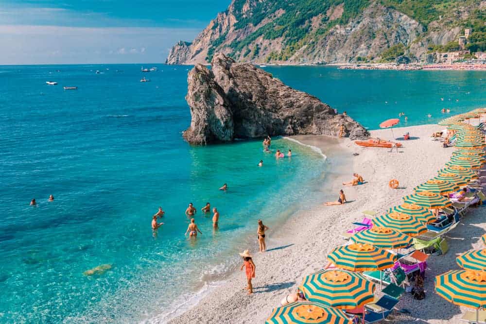 Striped beach umbrellas line the shore of Fegina Beach in Cinque Terre, where people swim and wade in aquamarine water.