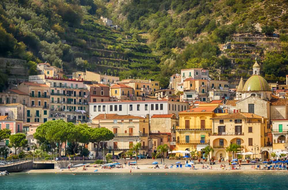 The yellow and brown buildings of Mairoi stand behind a sandy beach on the Amalfi Coast in Italy on a sunny day.