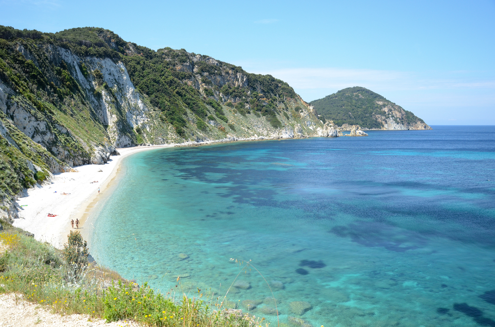 Green-covered stone cliffs flank a crescent-shaped sandy beach in Italy with bright blue water.