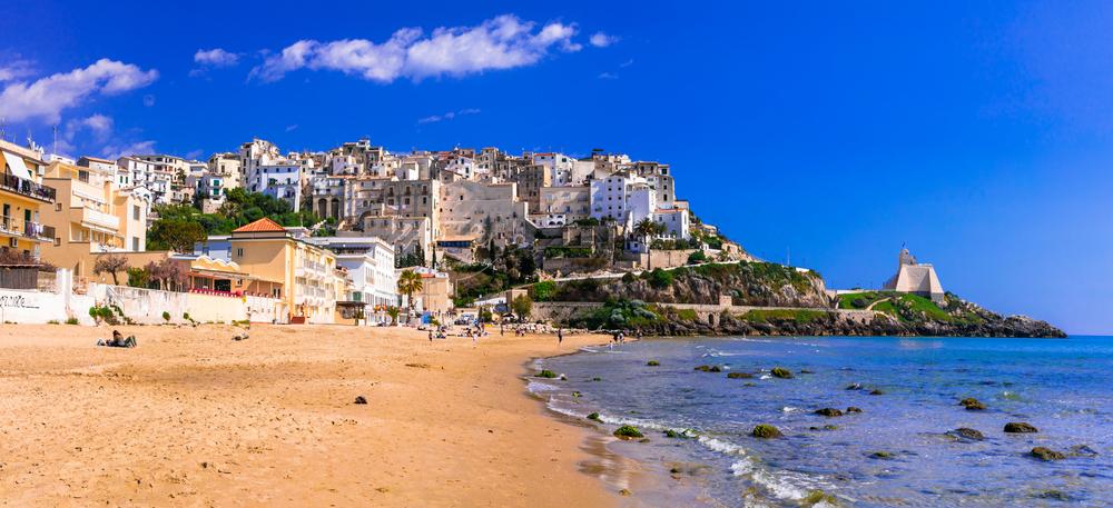 The town of Sperlonga sits on a hill, with the public beach in the foreground, on a bright, blue-sky day.
