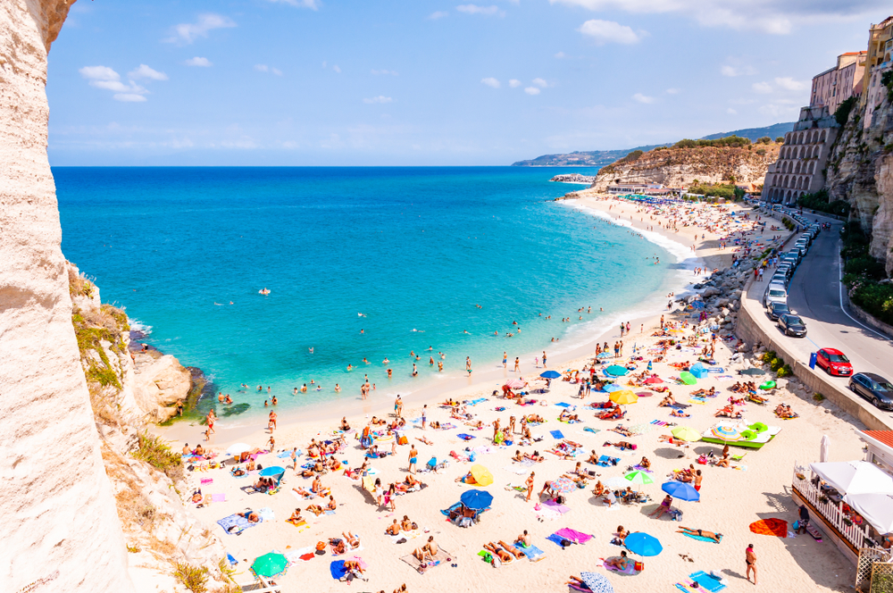 Colorful umbrellas and towels dot the Rotunda Beach in Tropea, with a narrow street running parallel to the bright blue water.