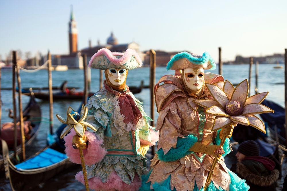 Two people dressed in colorful Carnevale costumes with masks near gondolas in Venice.