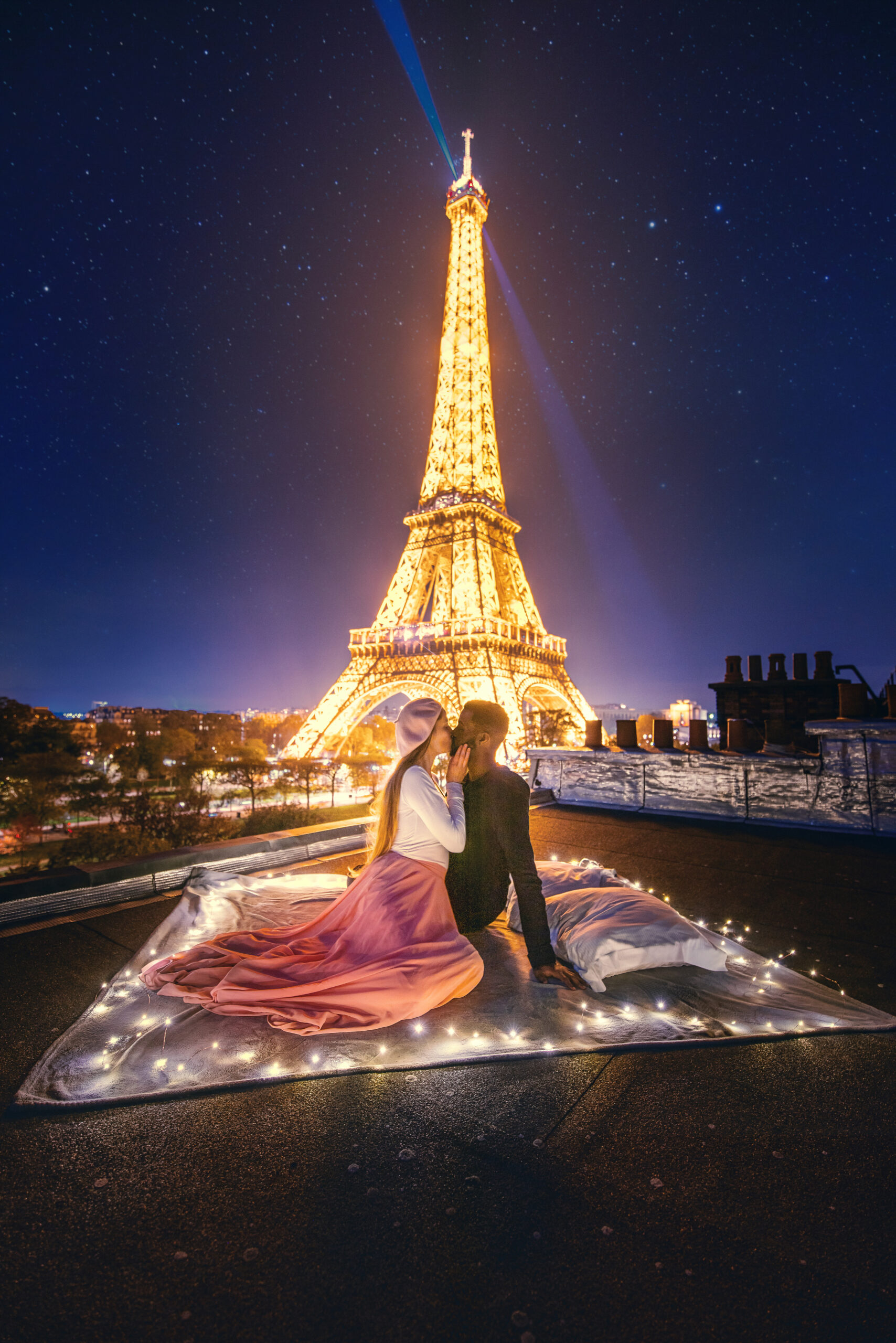 Couple sits kissing on a rooftop overlooking the lit up Eiffel Tower at night.