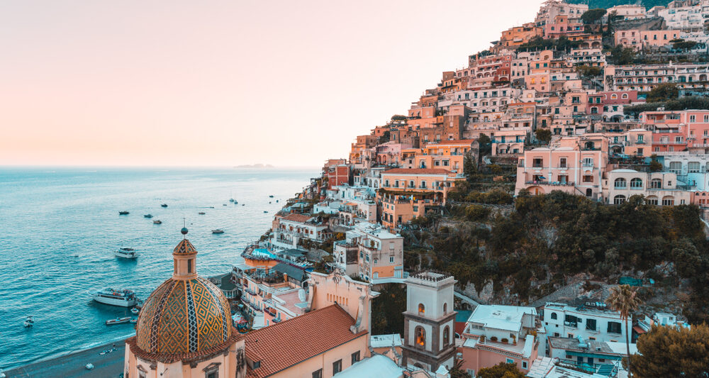 Rows of houses sit on a hill beside a calm blue sea along the Amalfi Coast at sunset