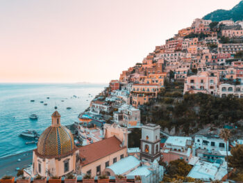 Rows of houses sit on a hill beside a calm blue sea along the Amalfi Coast at sunset