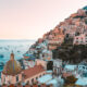 Rows of houses sit on a hill beside a calm blue sea along the Amalfi Coast at sunset