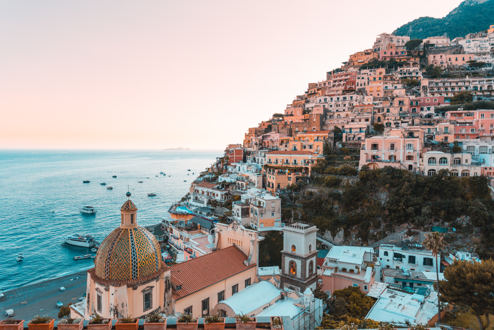Rows of houses sit on a hill beside a calm blue sea along the Amalfi Coast in Italy at sunset