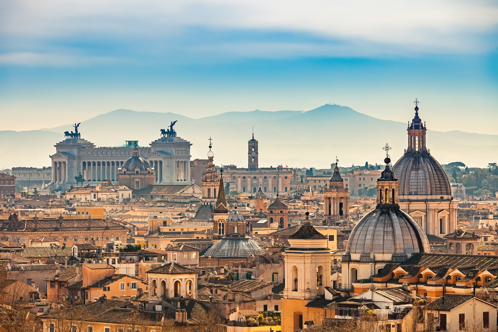 The spires and domes of the rooftops of Rome, with warm-colored buildings and hills in the distance.