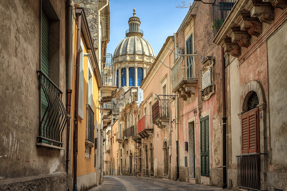 An empty street in Sicily (a popular place to visit in Italy in September), with brown two-storied buildings and the dome of a larger building in the distance