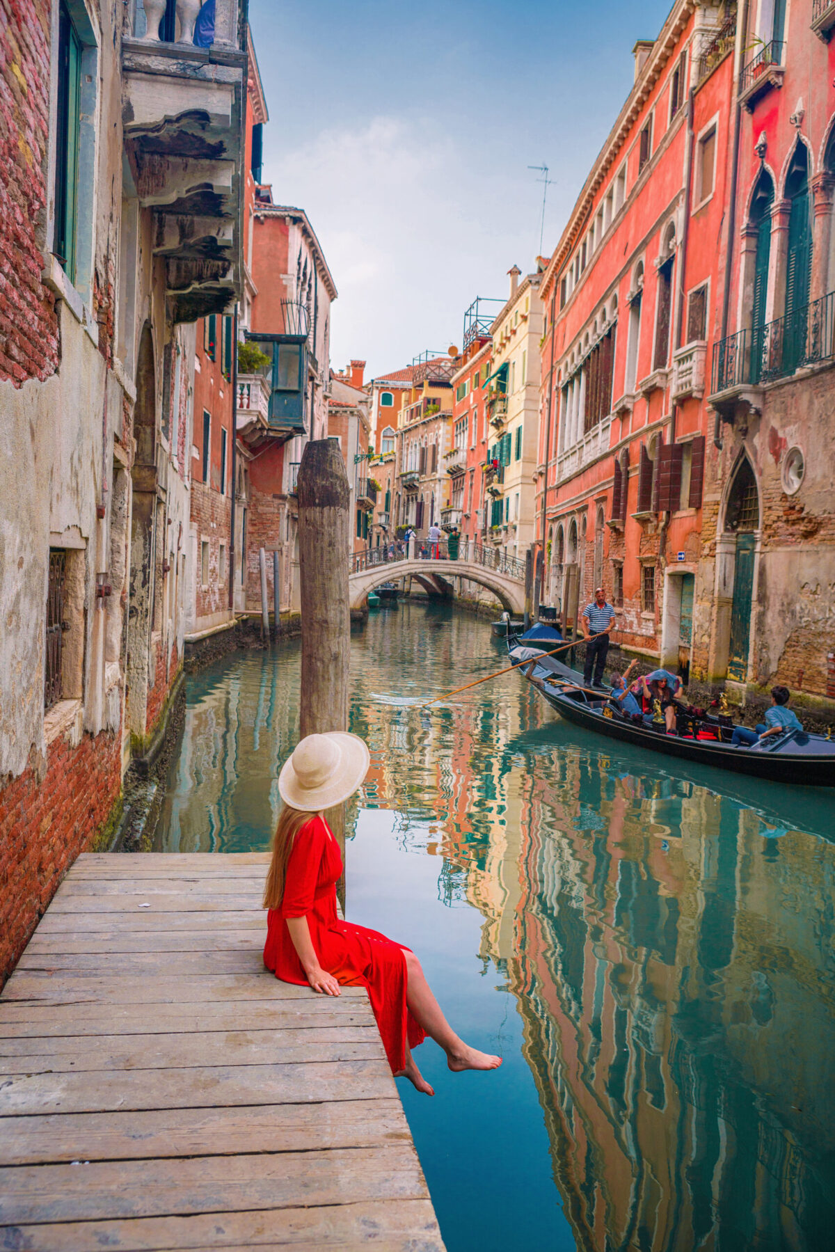 A woman with long hair and in a long, red dress, sits on a dock looking at a boat go by on a canal in Venice, Italy.