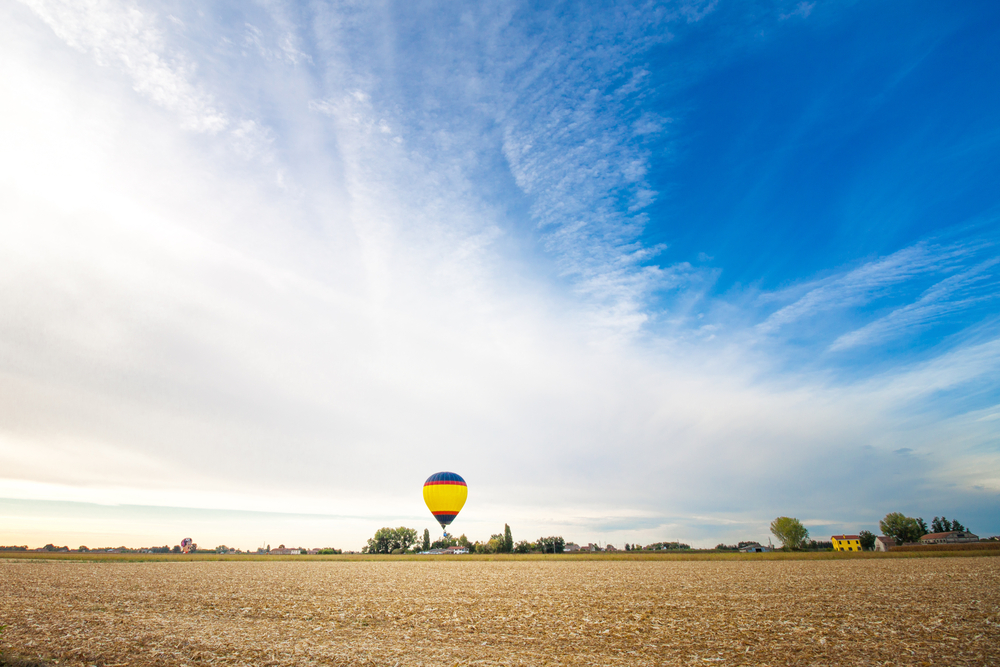 A yellow, red, and blue hot air balloon flies over a brown farm field in Italy in September as part of the festival in Ferrara.