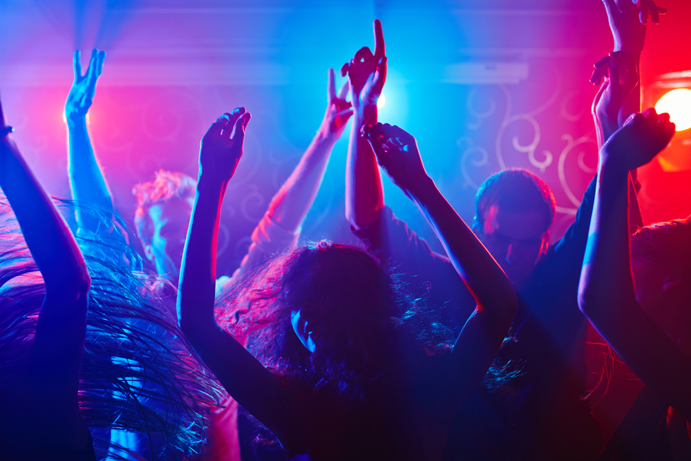 People with their hands in the air dance at a nightclub with blue and red lights shining.