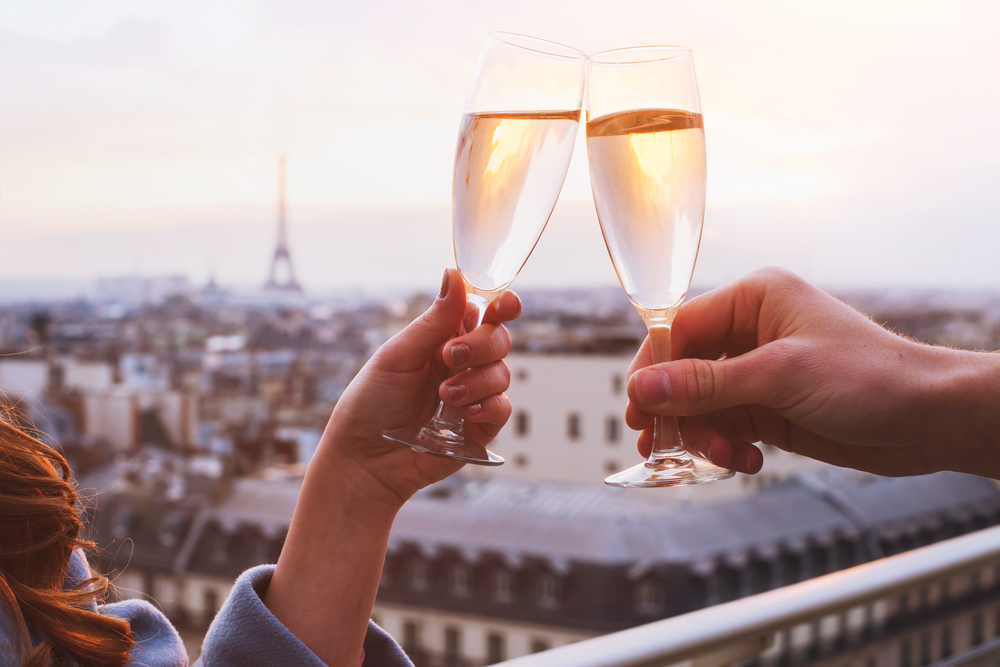 Close up of a couple clinking two champagne glasses with a view of a pink sunset over Paris.