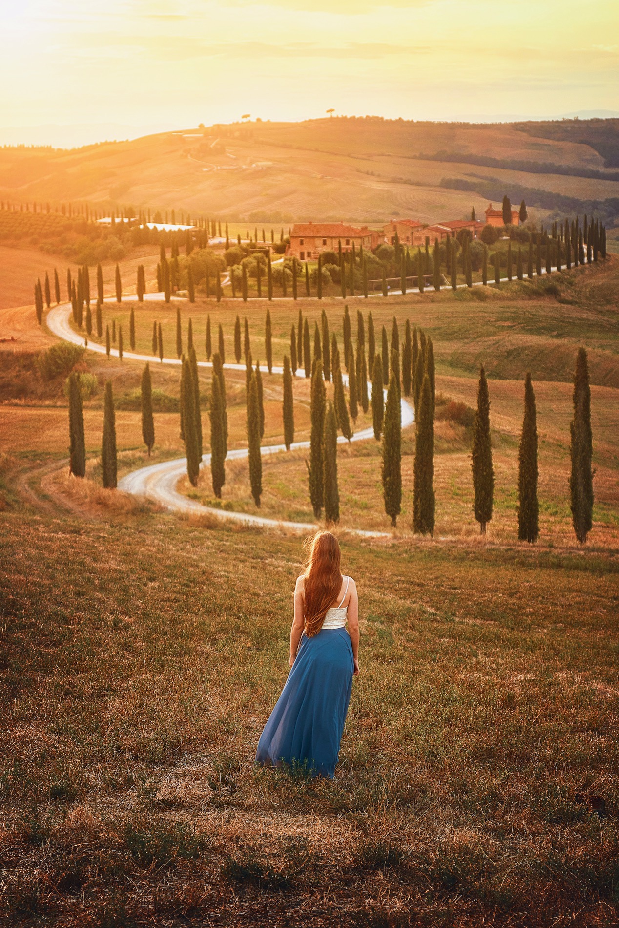 Woman in blue skirt stands overlooking the Tuscan landscape with iconic trees and a villa in the distance.