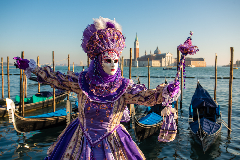 Woman in an elaborate, purple dress and mask in front of gondolas in Venice.