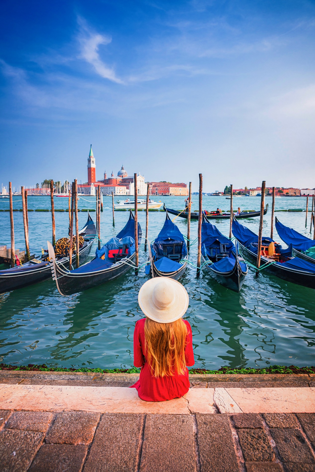Woman in red and sun hat sits on the edge of the lagoon near docked gondolas in Venice.