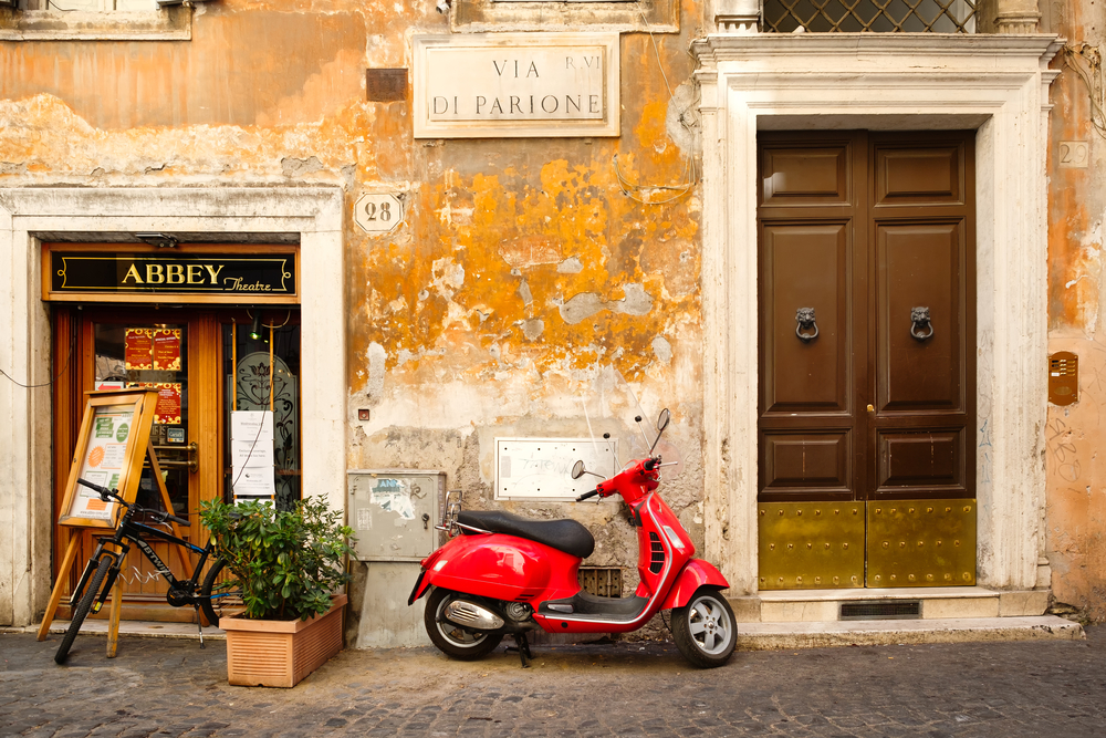 Red Vespa leaning against a textured wall on a street in Rome.