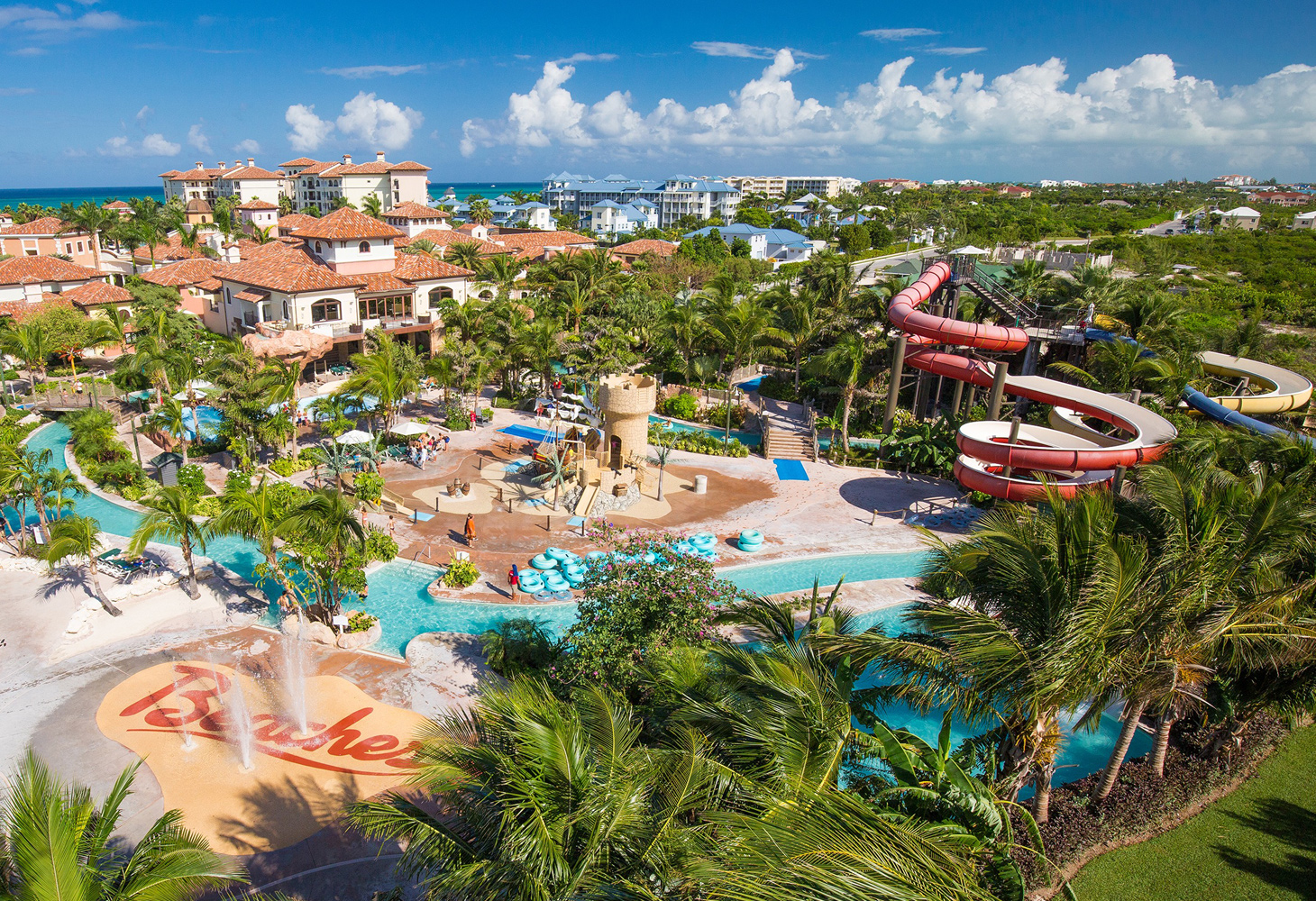 An aerial view of a large resort with pools, a sandcastle style playground, and water slides surrounded by palm trees