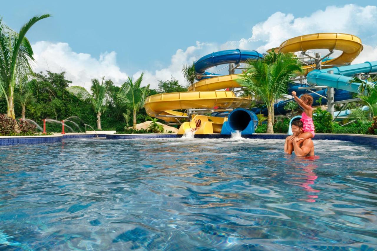A dad and his daughter playing in a pool with two waterslides attached to it surrounded by palm trees at one of the best all inclusive resorts for families